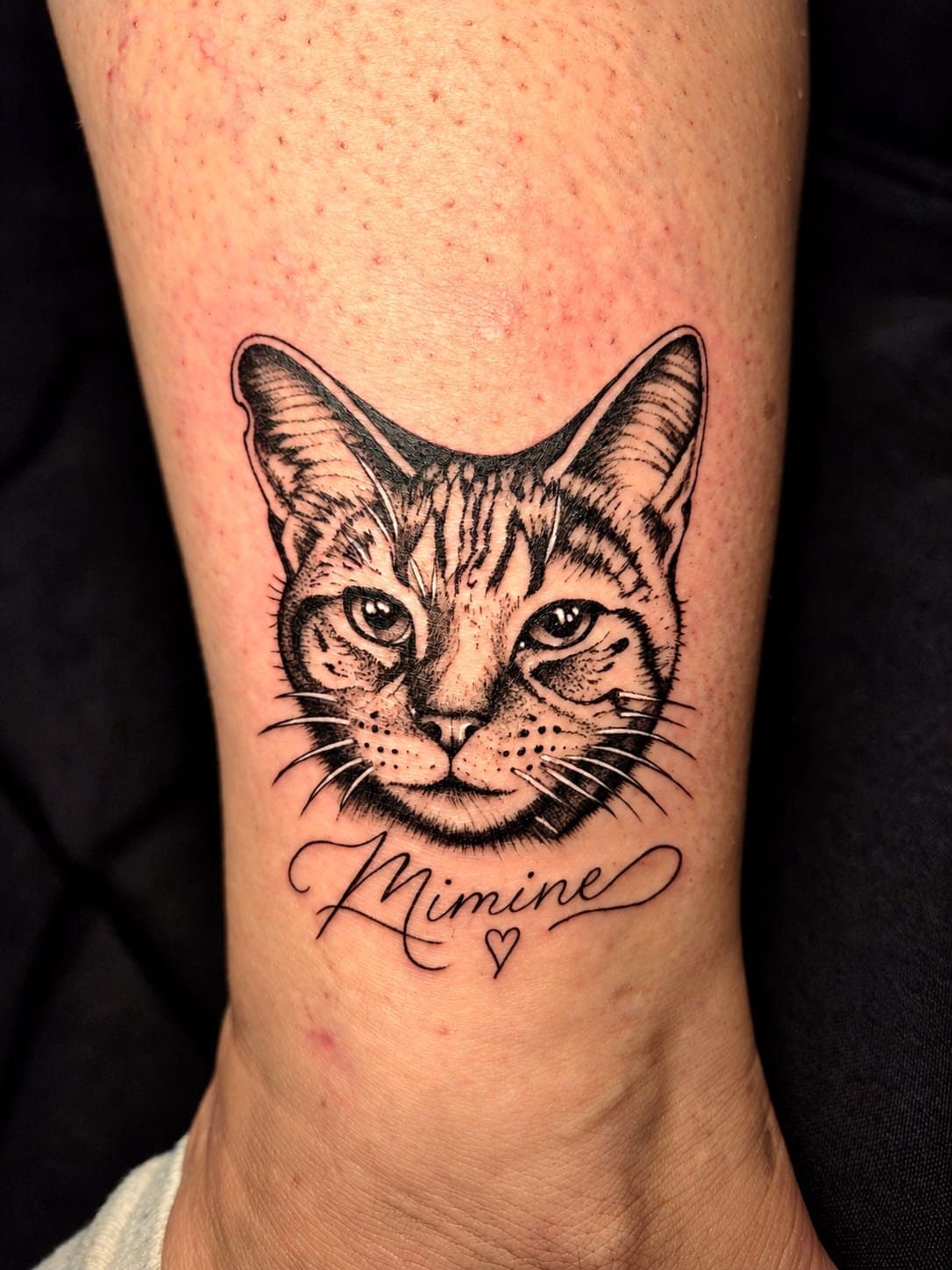 Portrait de chat tigré tatoué en micro réalisme sur la cheville, avec le prénom Mimine en lettering fin