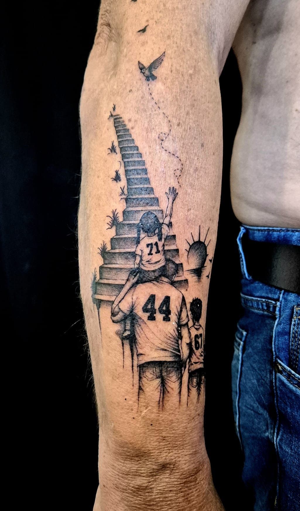 Père portant sa fille sur les épaules au pied d'un escalier vers le ciel, maillots numérotés, oiseau et soleil levant, tatoué en noir et gris sur l'avant-bras