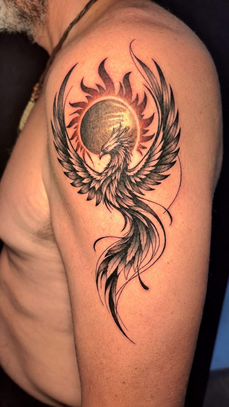 Phénix aux ailes déployées devant un soleil, tatoué en ligne fine et dotwork sur le bras, à Gruissan